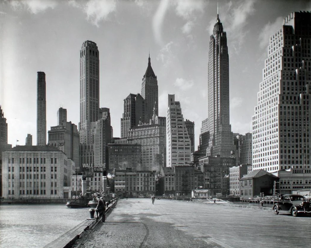 A black and white 1930s photo of the downtown skyline from an East River pier, tall Art Deco towers with spires rising behind low waterfront buildings, a man on the pier and a vintage car at right