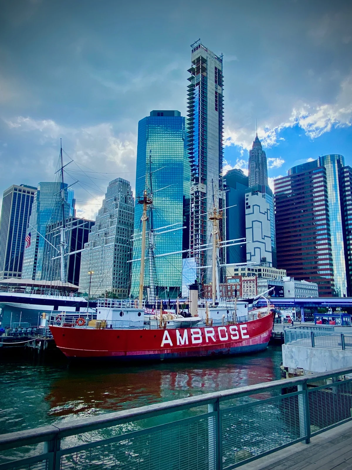 A red lightship named Ambrose docked beside a pier, with tall ship masts and glass towers of the Financial District behind it under a dramatic sky