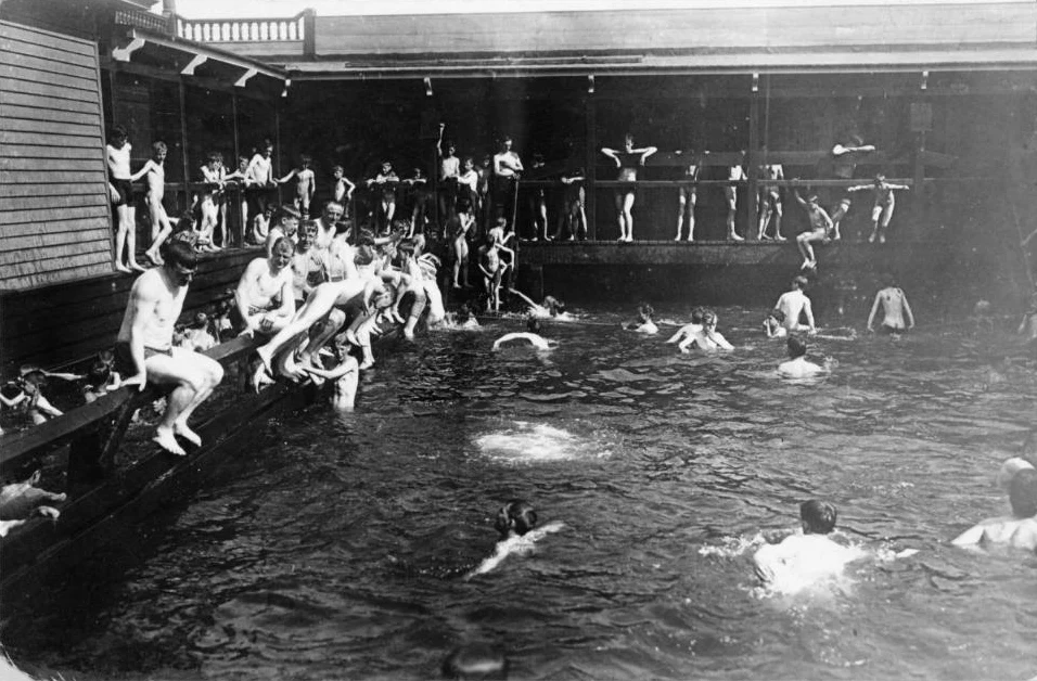 A black and white 1908 photo of crowds of boys swimming and diving in a wooden floating bath