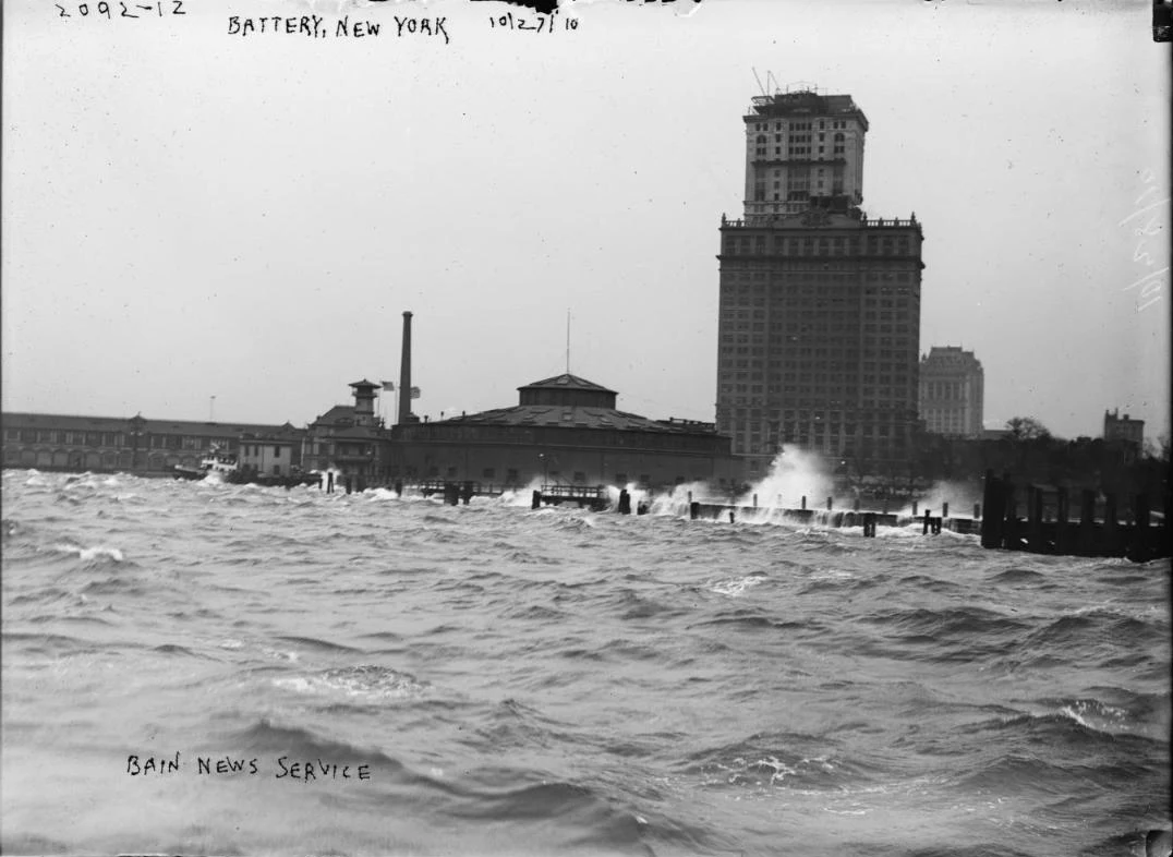 A black and white 1910 photo of storm waves breaking over the Battery seawall, with low waterfront buildings and a tall office tower behind