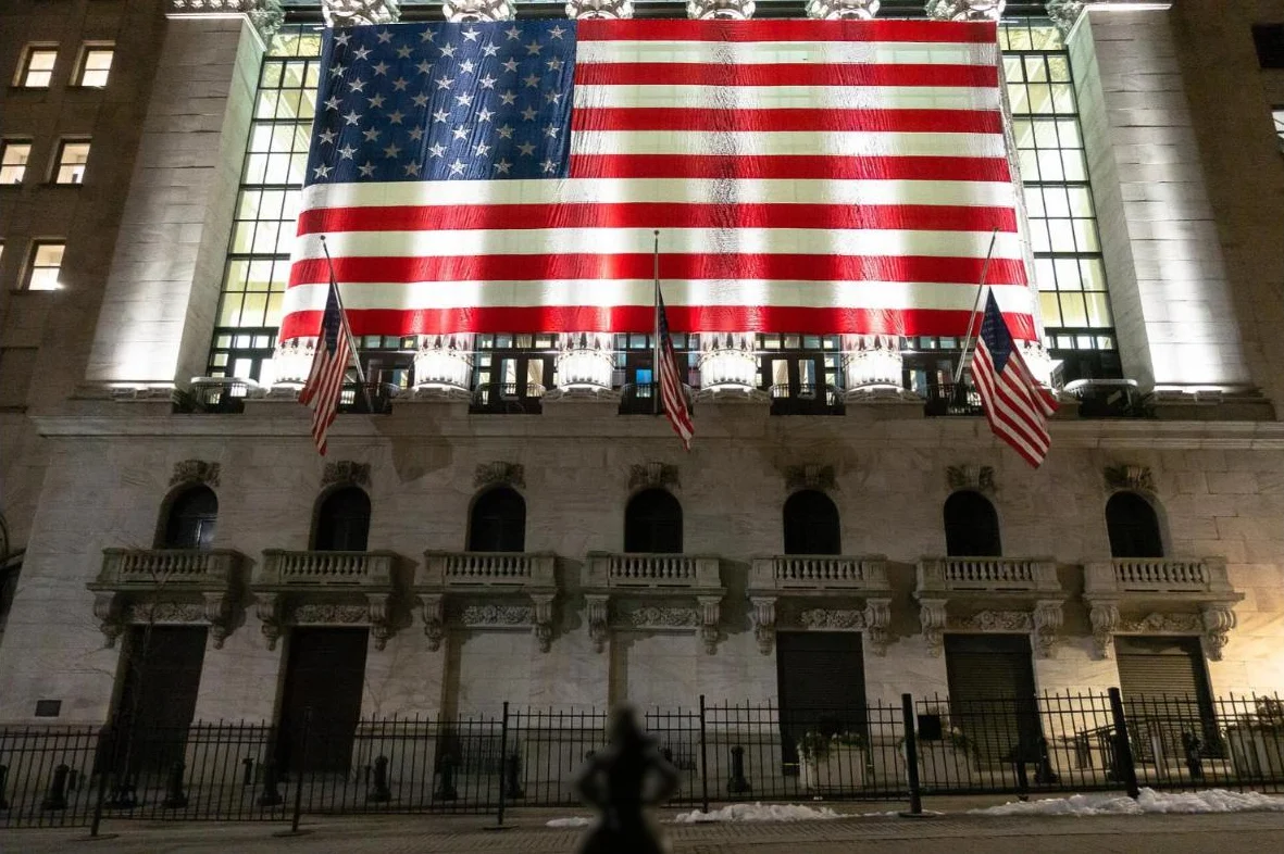 The Stock Exchange lit at night with a giant American flag, and the small silhouette of the Fearless Girl statue in front