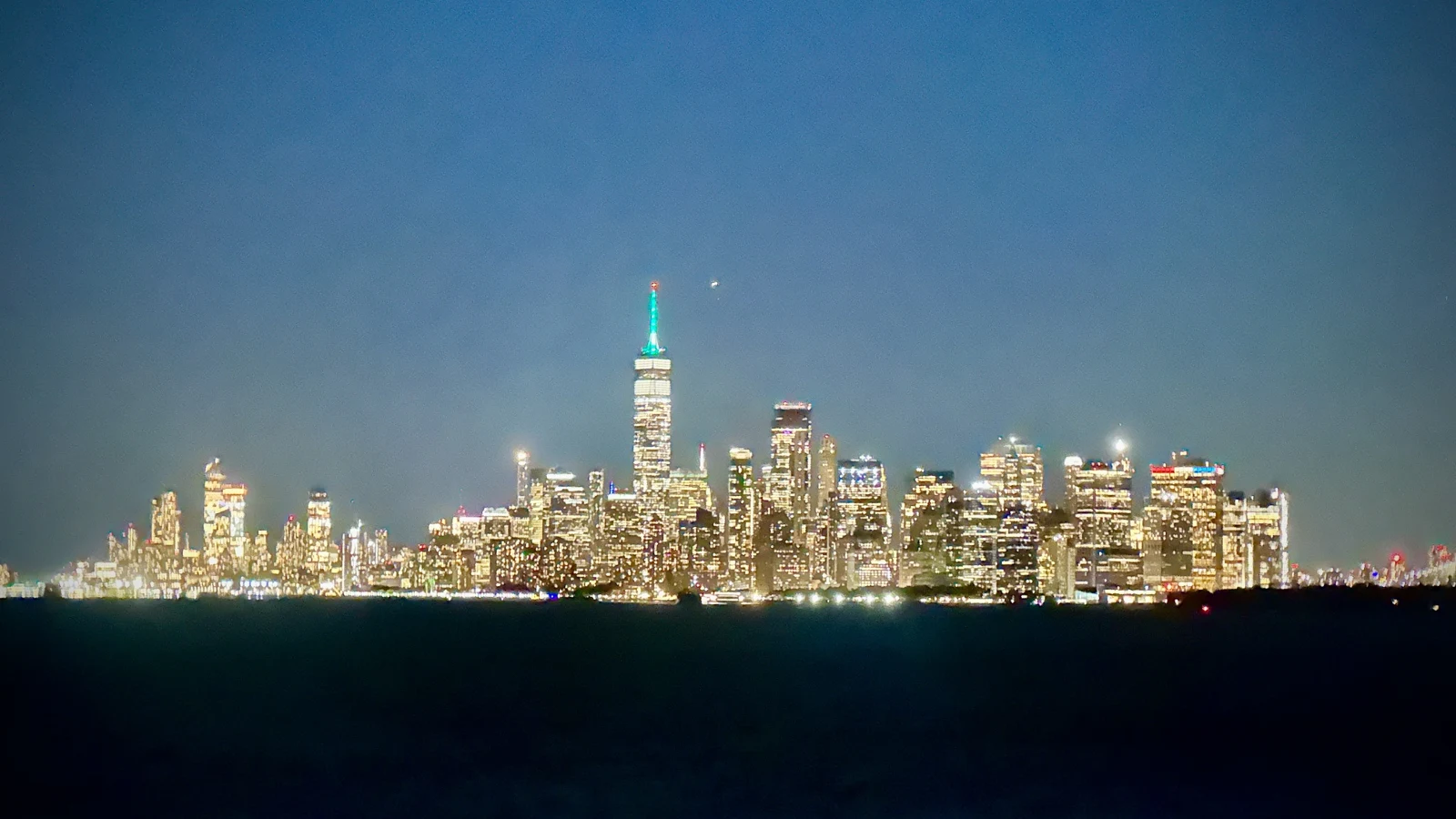The Lower Manhattan skyline lit up at night across dark harbor water, One World Trade Center glowing at the center under a deep blue sky