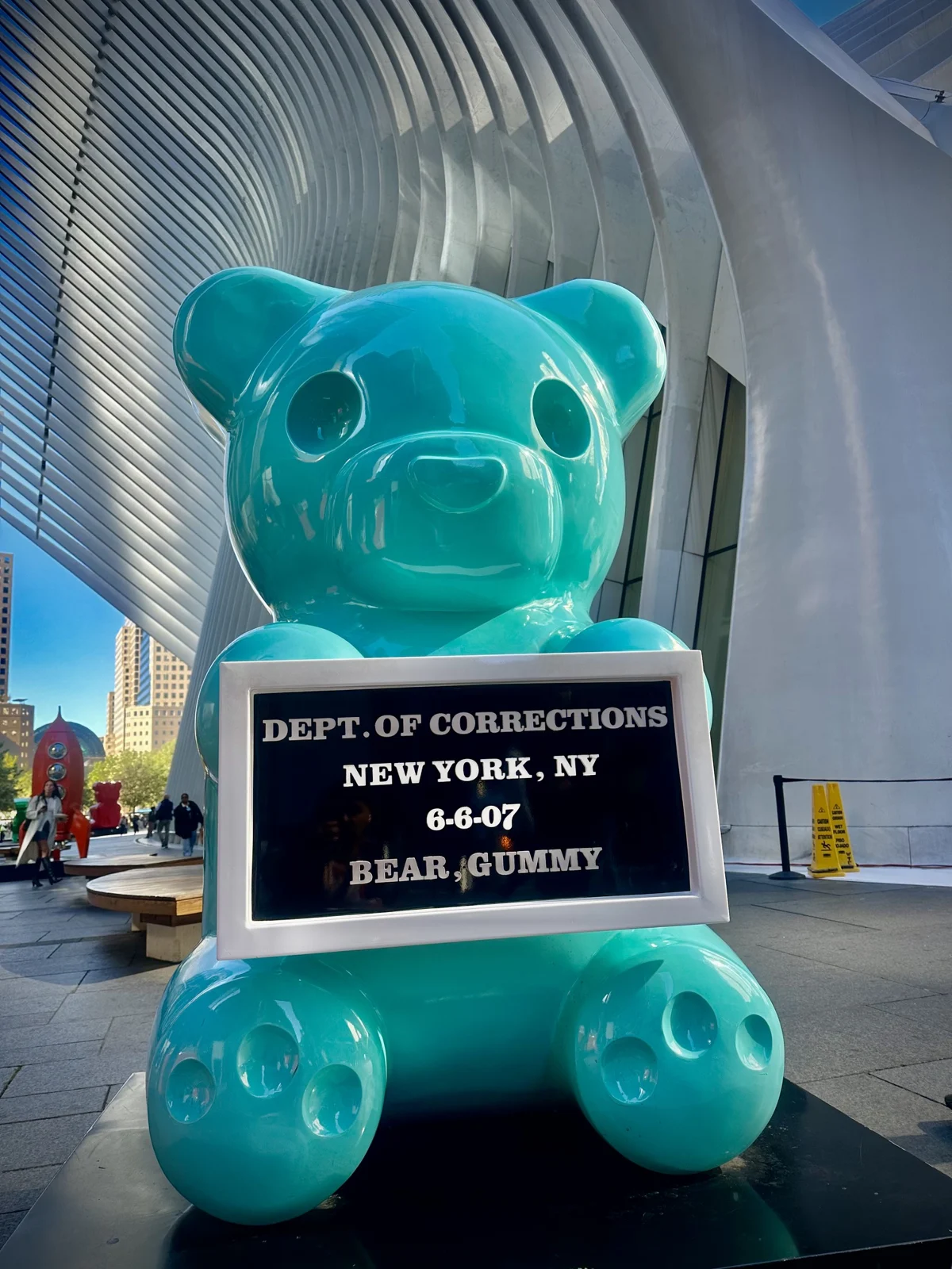A giant glossy turquoise gummy bear sculpture holding a booking board that reads Dept. of Corrections, New York, NY, Bear, Gummy, under the white ribs of the Oculus, with a red rocket sculpture behind