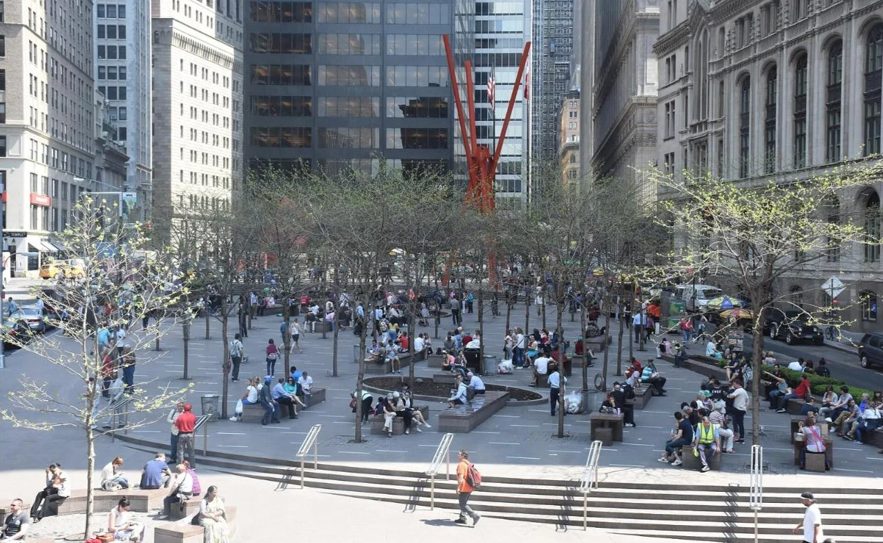 A tall red steel sculpture above a busy tree-lined plaza full of people
