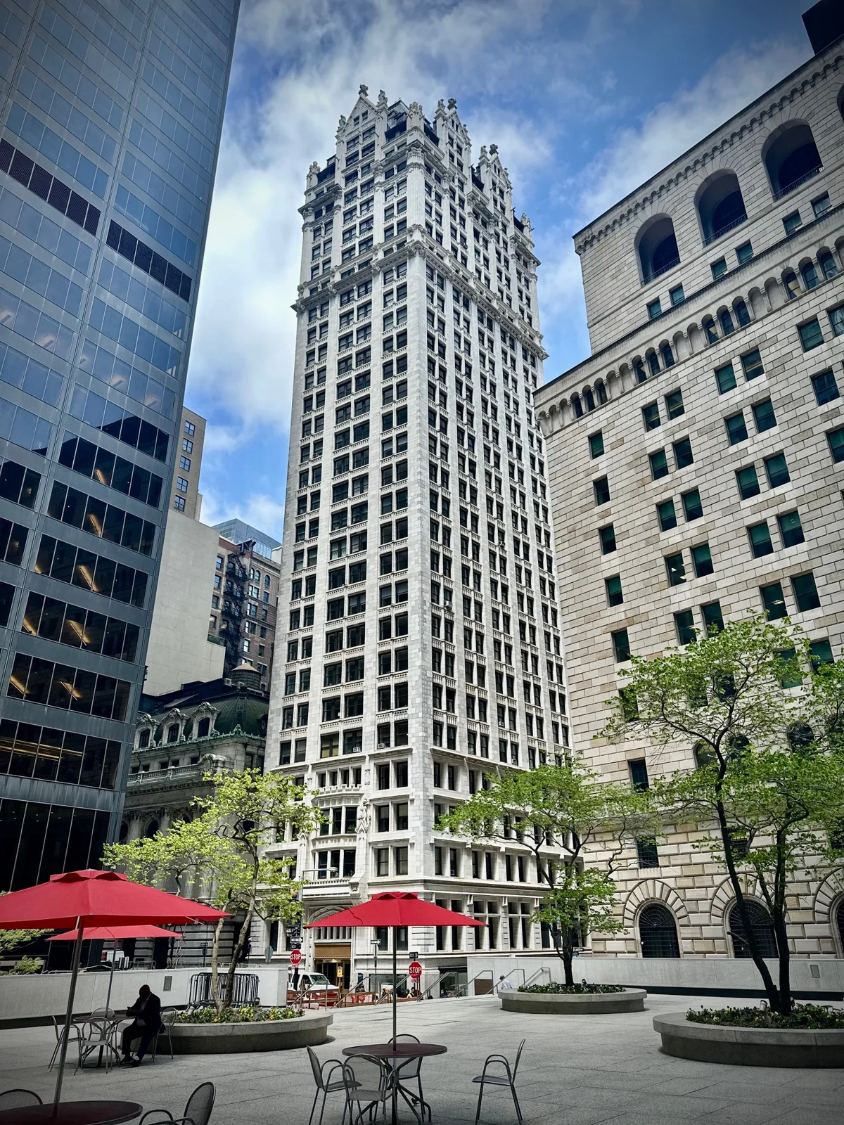 A slender white Gothic terra-cotta skyscraper rising above a plaza with red umbrellas and trees, between a glass tower and a heavy stone bank building