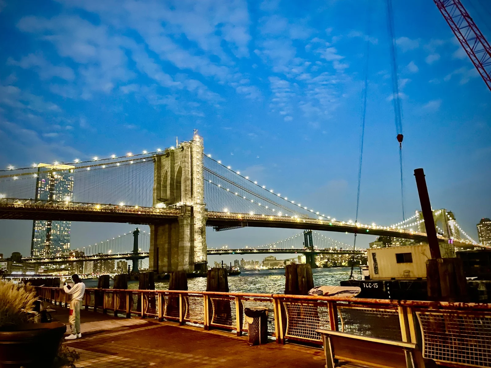 The Brooklyn Bridge lit with strings of lights at dusk, the Manhattan Bridge behind it, seen from a waterfront promenade with a railing and benches