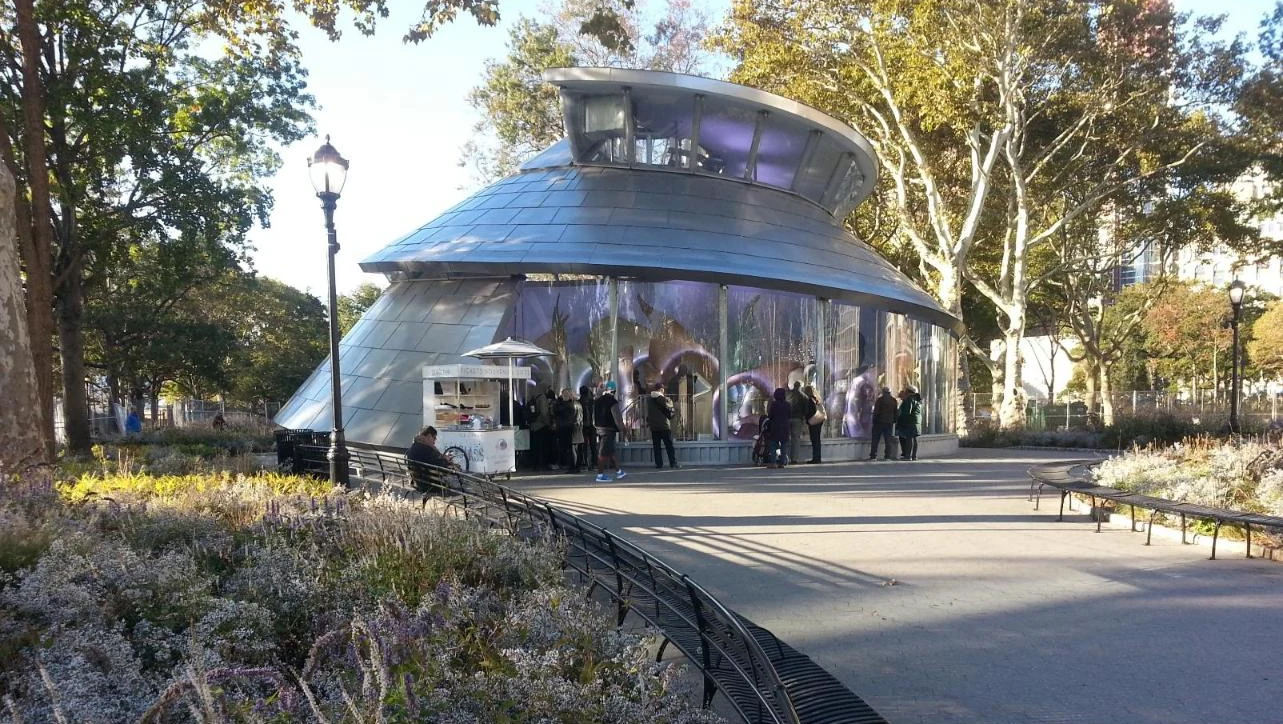 A round pavilion with a curved metal roof and glowing glass walls, surrounded by trees and flowering garden beds