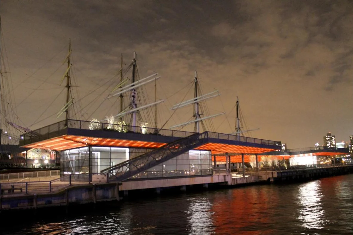 A two-level pier lit orange at night, reflected in the river, with the masts of tall ships behind