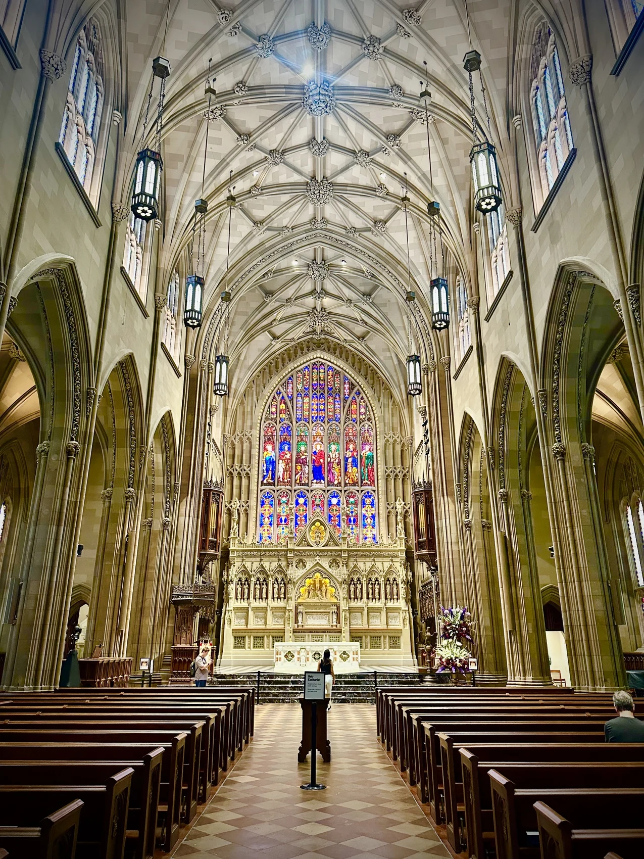 Looking down the center aisle of Trinity Church past wooden pews toward a carved white altar screen and a tall stained-glass window, under a ribbed and vaulted ceiling