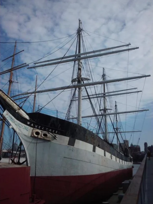 The bow and tall masts of a historic iron sailing ship docked at a pier under a cloudy blue sky