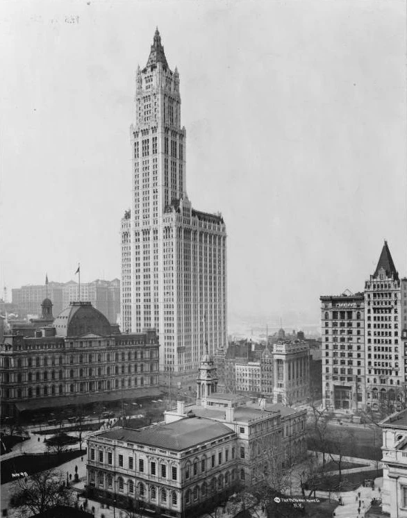 A black and white 1913 photo of the new Woolworth Building&rsquo;s Gothic tower rising behind City Hall and the old domed post office, with bare trees in City Hall Park