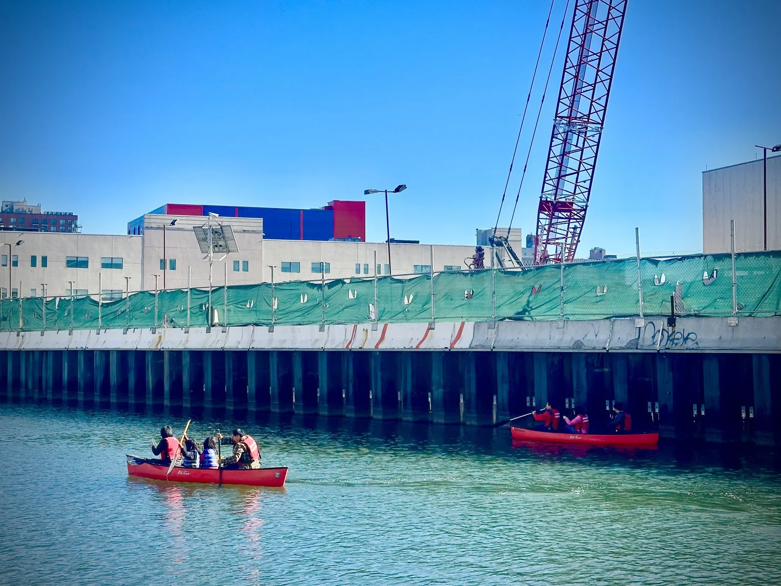 Two red canoes full of paddlers in life vests gliding along the green water of the Gowanus Canal beside a bulkhead, with a red crane and fenced construction site above and a red and blue building beyond