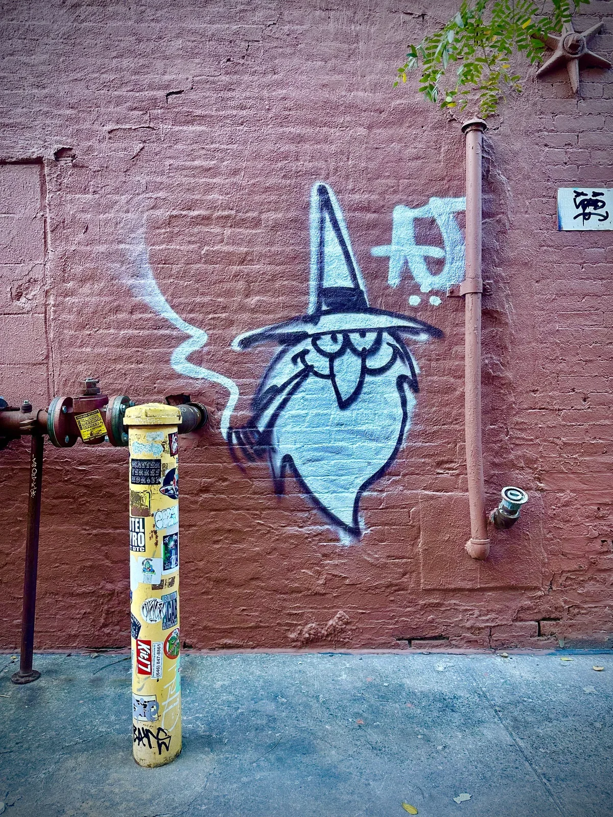 A spray-painted cartoon wizard in a pointed hat, puffing a pipe, on a rust-red painted brick wall beside a sticker-covered yellow standpipe and old iron pipes