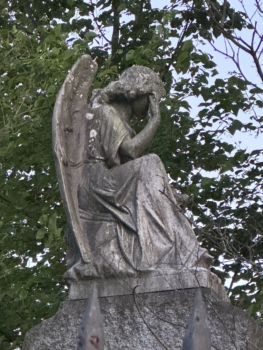 A weathered marble angel, head bowed in grief, atop a granite monument framed by green leaves