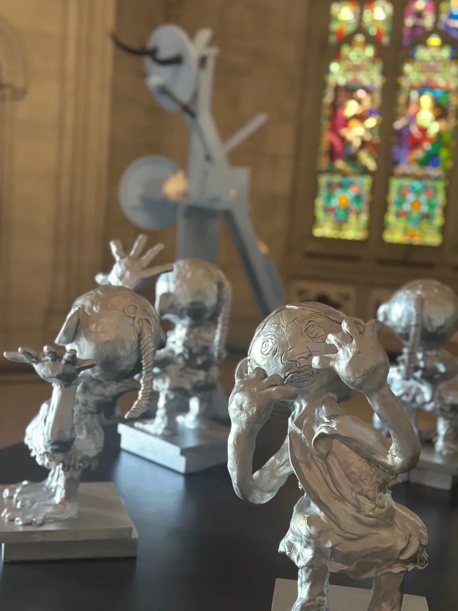 Silver sculpted figures on a dark table in the Chapel, with a pale blue sculpture and glowing stained glass behind them