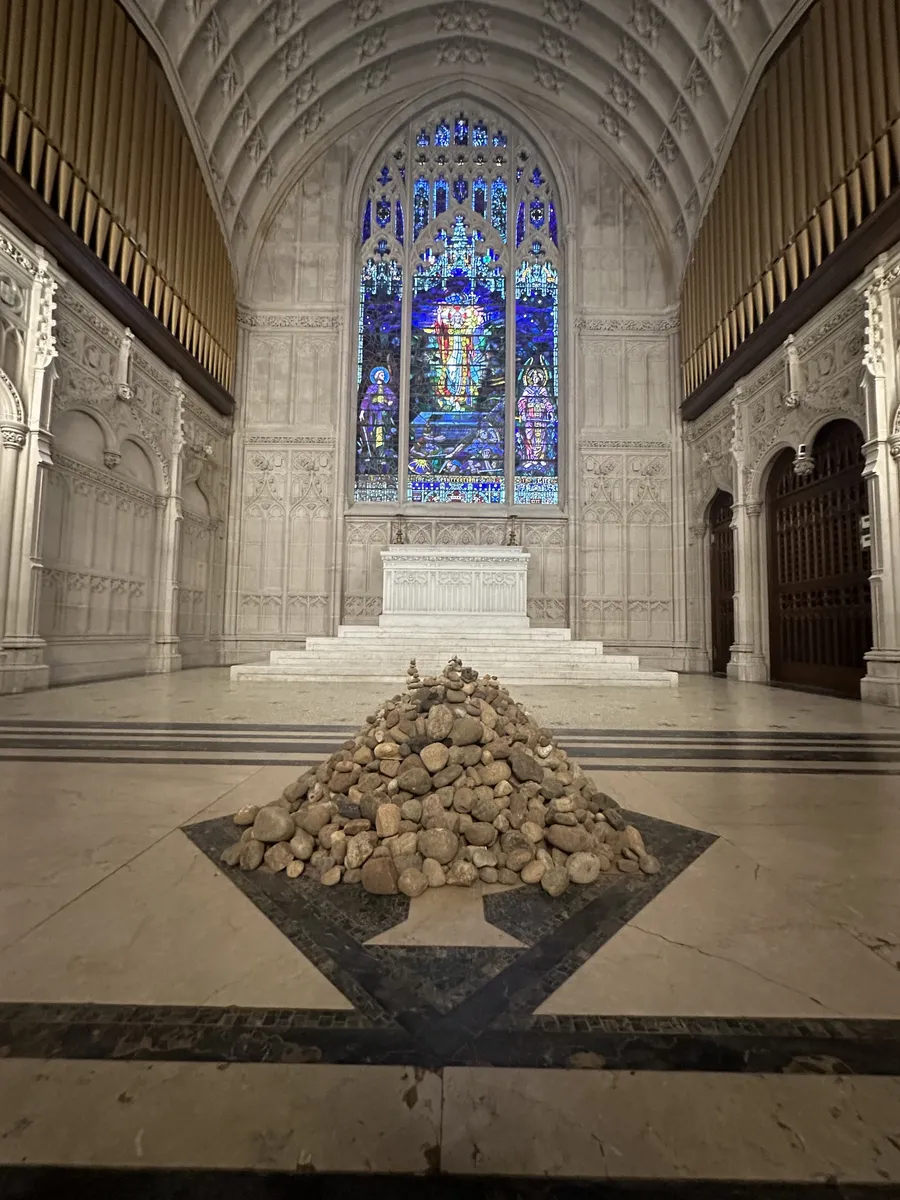 A mound of river stones on the marble floor of the Chapel, beneath a blue stained-glass window and the altar