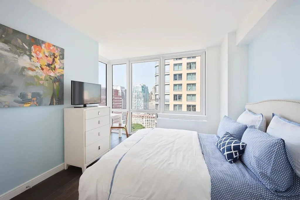 A bedroom with a blue and white bed and tall windows over the towers of Downtown Brooklyn